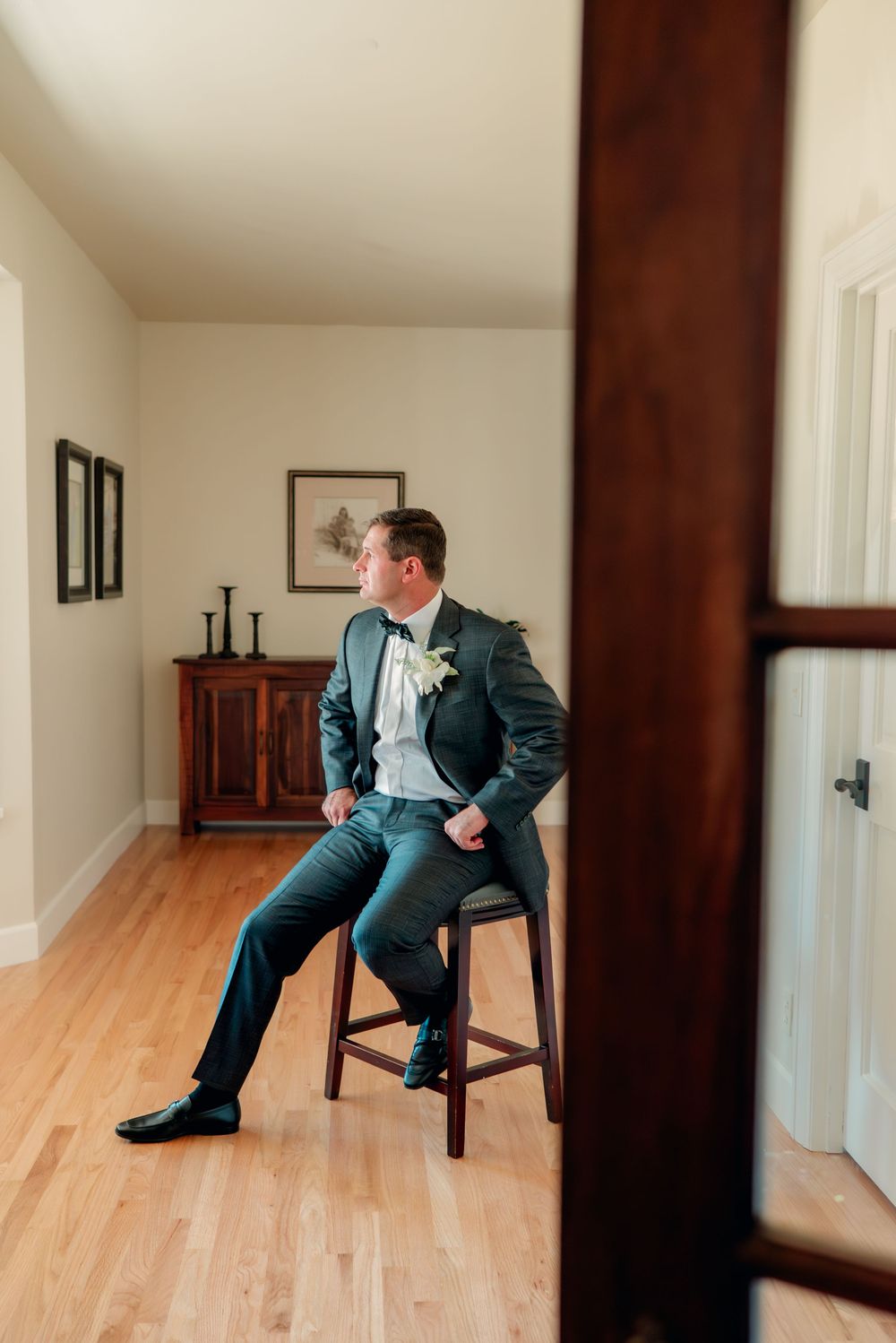 Figure in dark suit sits contemplatively on wooden chair in minimalist room with hardwood floors.