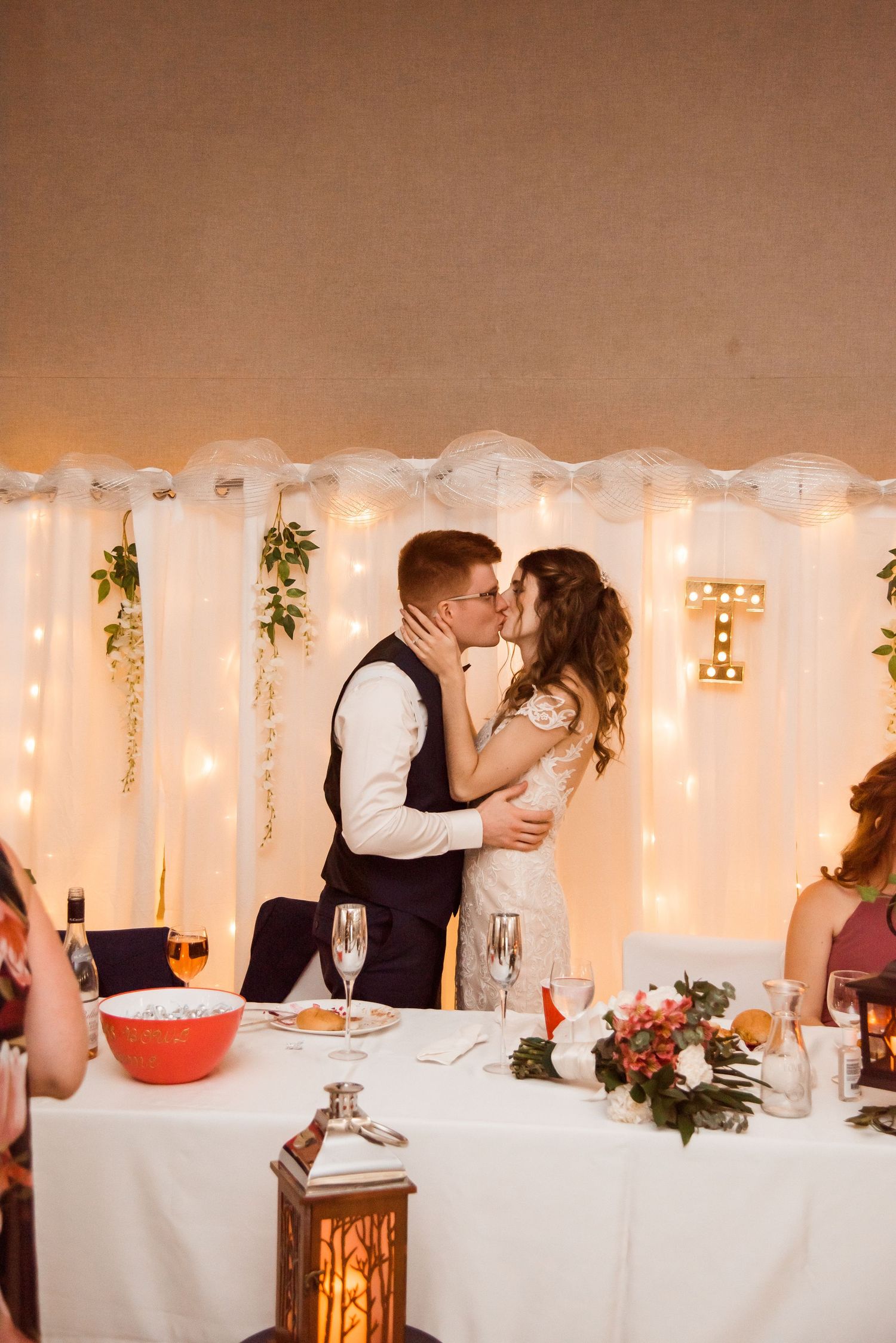 A couple shares a romantic kiss at their wedding reception with white decor and string lights in the background.