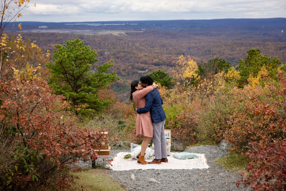 Couple embracing during outdoor fall engagement proposal with colorful autumn foliage and picnic setup.