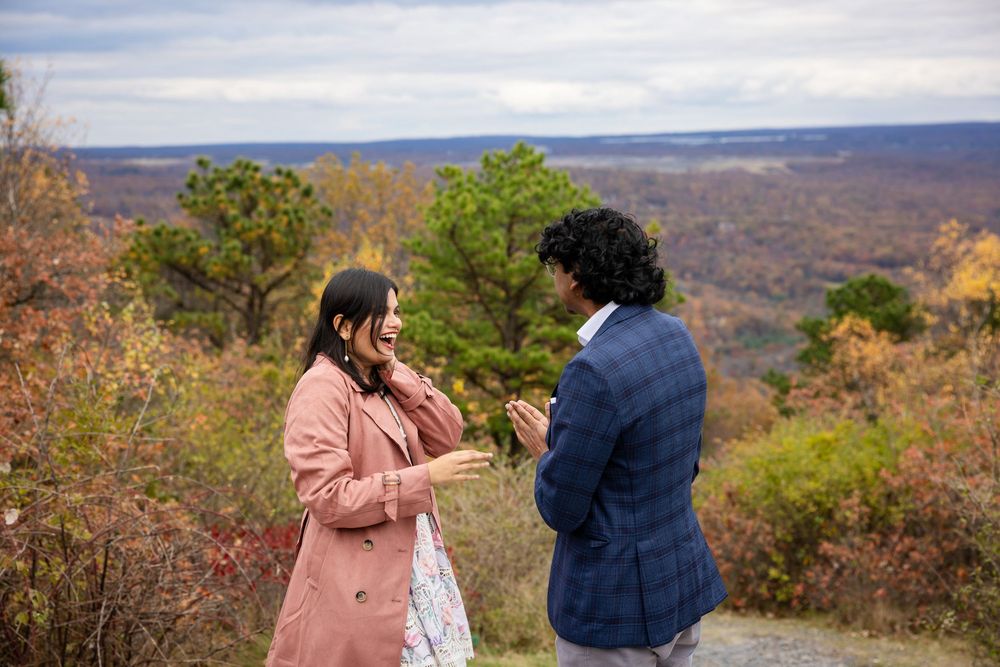 Couple sharing romantic proposal moment on scenic overlook with autumn foliage backdrop.