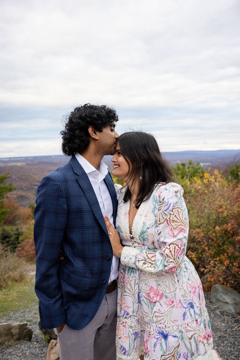 Man in blue plaid blazer kissing woman in floral dress on mountain overlook.