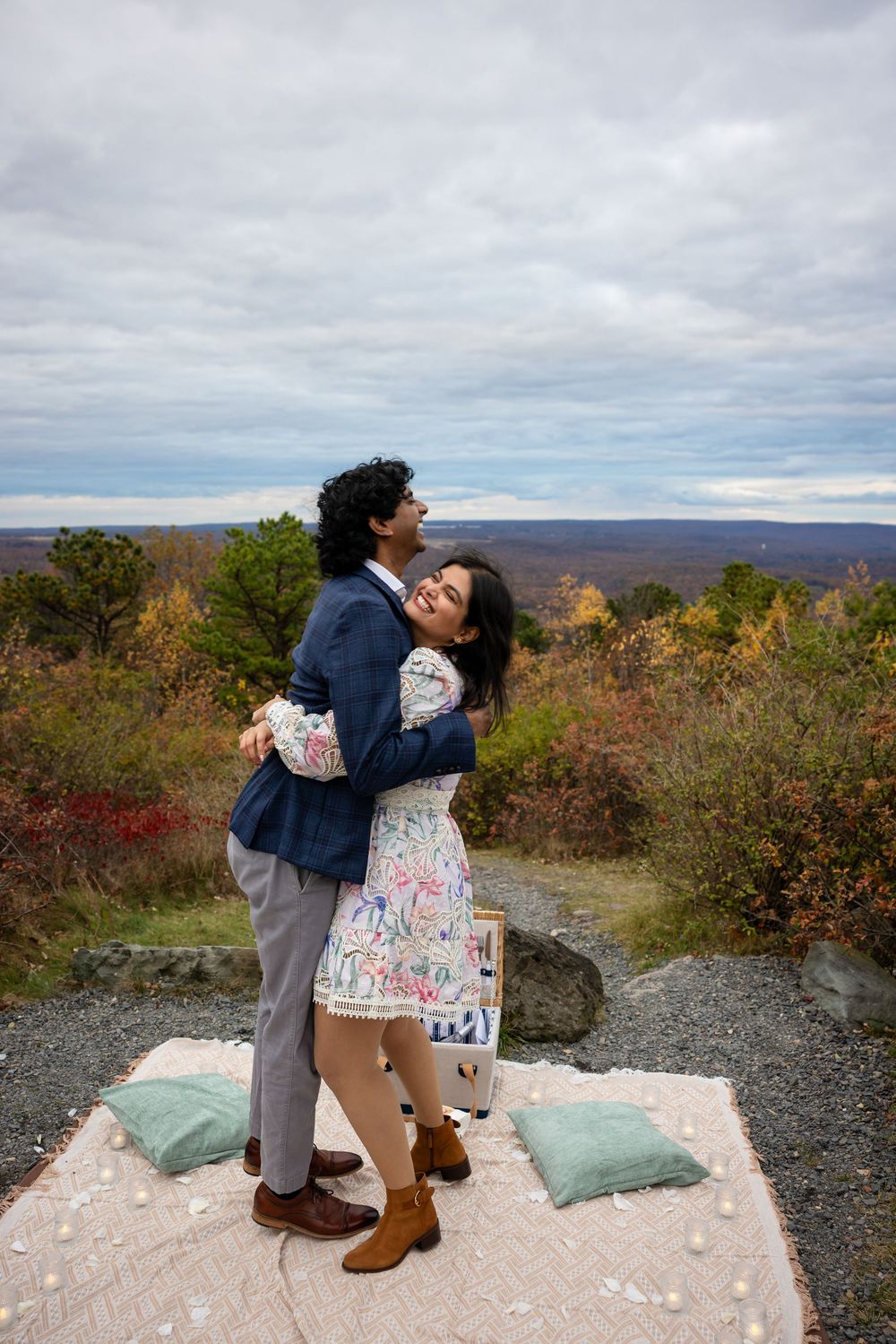 Couple embracing joyfully on blanket with colorful autumn mountain landscape behind them.