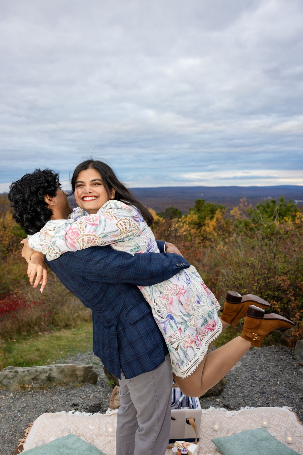 Man lifting smiling woman in floral dress on scenic autumn overlook outdoors.