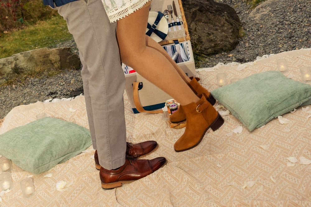 Close-up of couple's feet, man in brown dress shoes and woman in tan ankle boots on a picnic blanket outdoors.