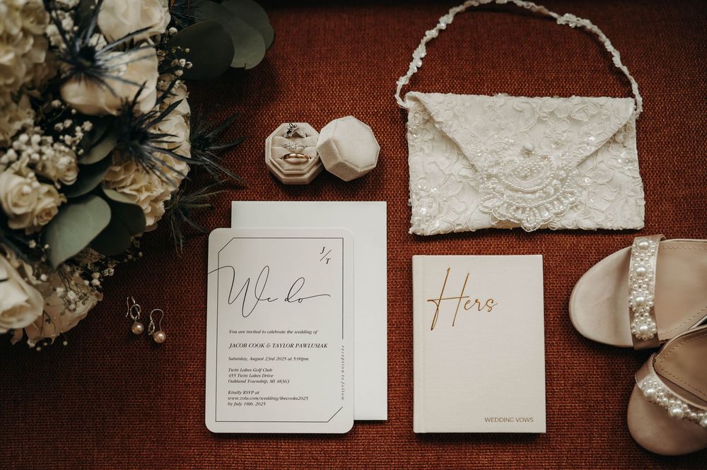 Elegant wedding flat lay featuring invitations, a lace clutch, pearl earrings, bridal shoes, and a ring box on red fabric.