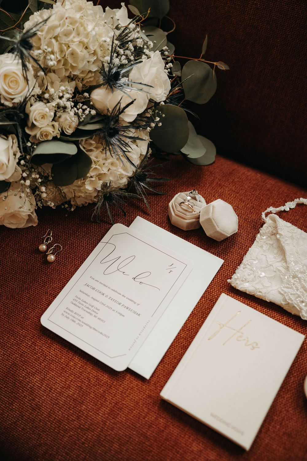 Wedding flat lay with white rose bouquet, elegant invitations, velvet ring box, and lace clutch on a red surface.