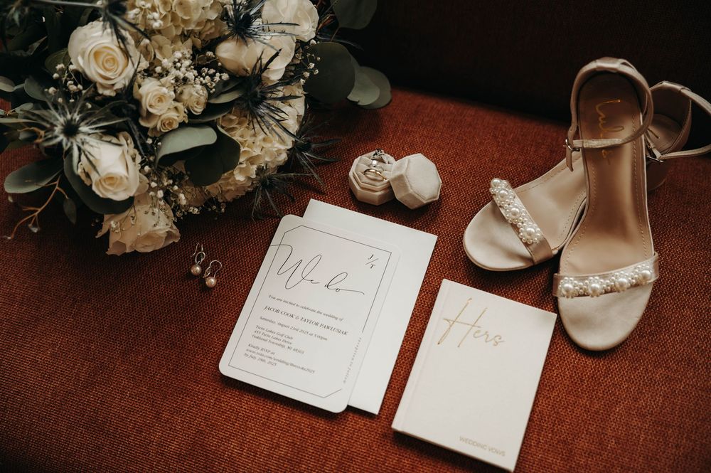 Bridal details flat lay with white rose bouquet, pearl-adorned heels, wedding invitations, and a ring box on red fabric.