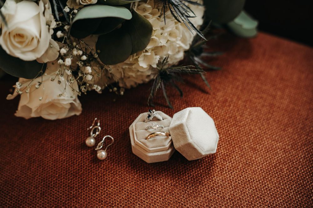 Close-up of wedding rings in a white velvet ring box beside pearl drop earrings and a white rose bridal bouquet.