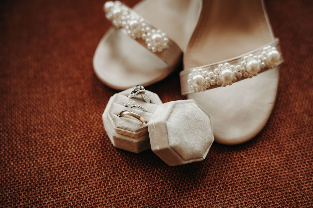 Wedding rings displayed in a white velvet ring box resting against pearl-embellished bridal heels on a rust-colored surface.