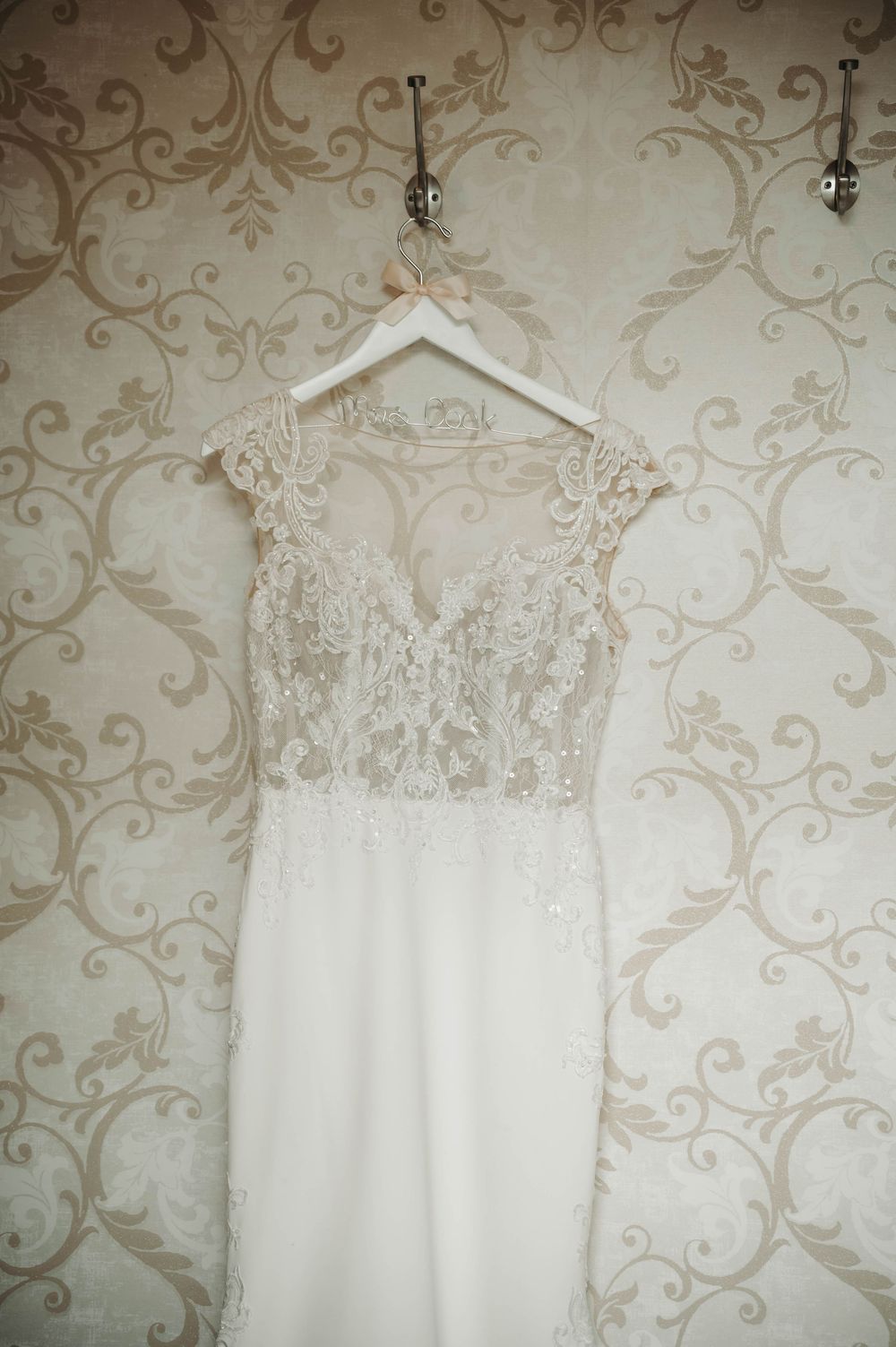 Elegant lace wedding gown with cap sleeves and V-neckline displayed on a white hanger against floral wallpaper.