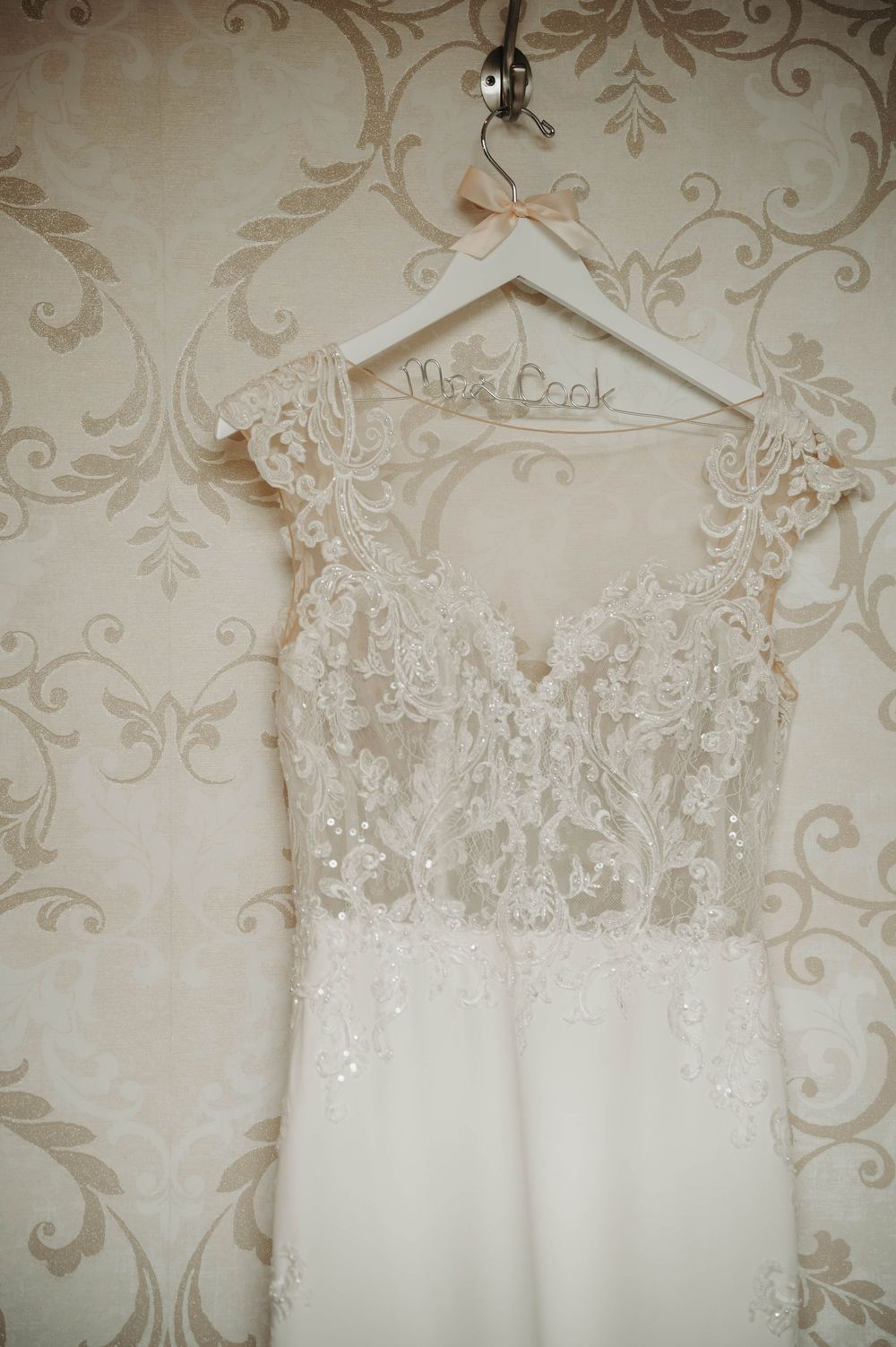 Lace wedding gown with cap sleeves hanging on a personalized Mrs. Cook hanger against vintage floral wallpaper.