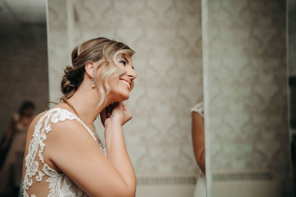 Smiling bride in lace wedding dress admiring her reflection in a mirror while touching her necklace.