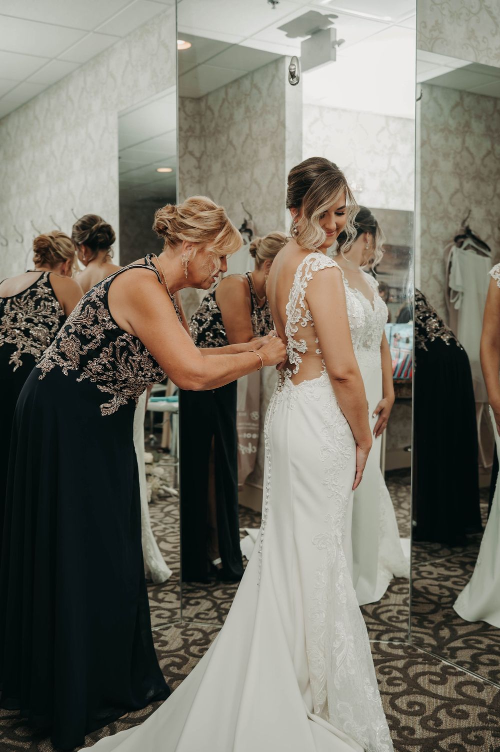 Woman helping bride button up elegant lace back wedding dress in a bridal suite mirror.