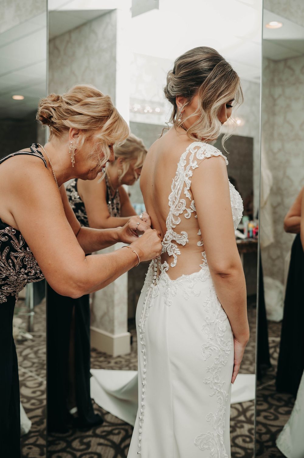 Close-up of woman fastening intricate lace back buttons on bride's fitted wedding gown.