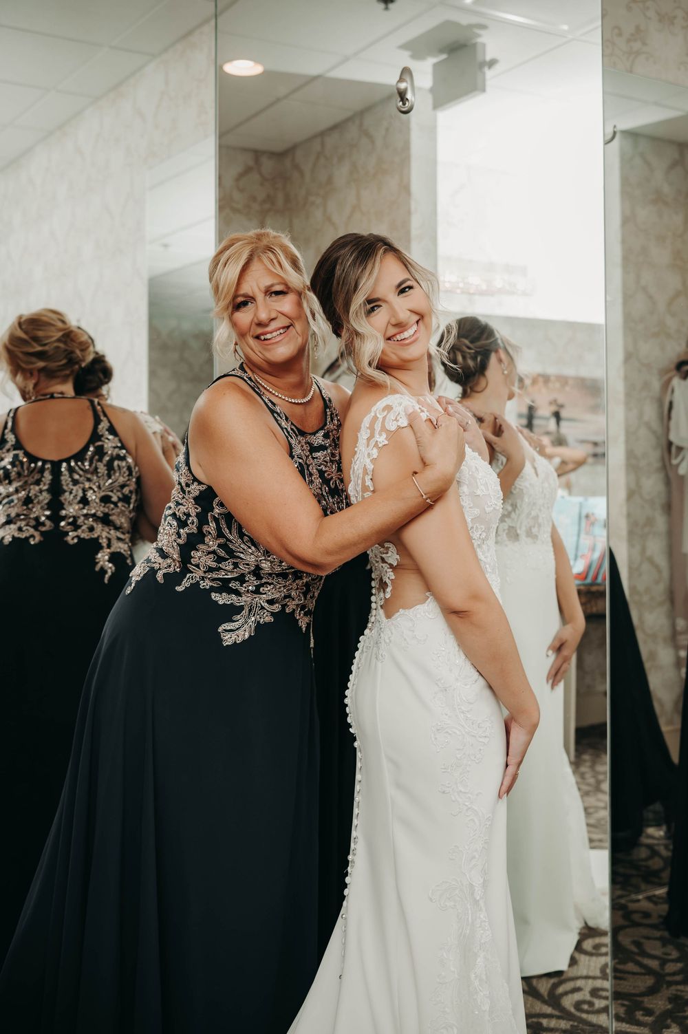 Mother of the bride embracing smiling bride in fitted lace wedding dress in bridal suite.