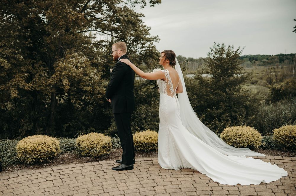 Bride surprising groom with first look moment on brick patio surrounded by lush greenery.