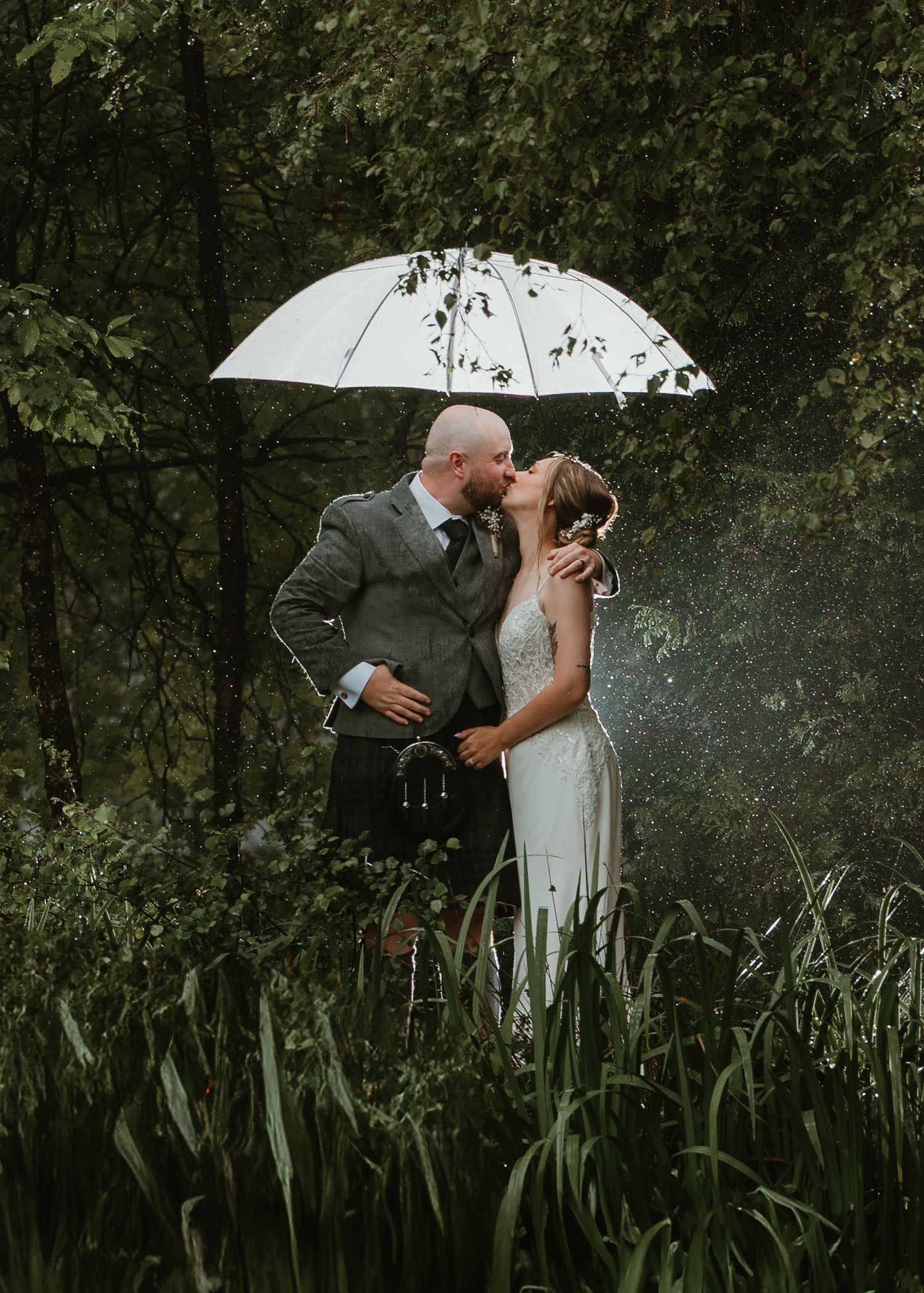 A romantic couple shares a tender kiss in a lush garden setting with tall grass and trees creating a dreamy atmosphere.