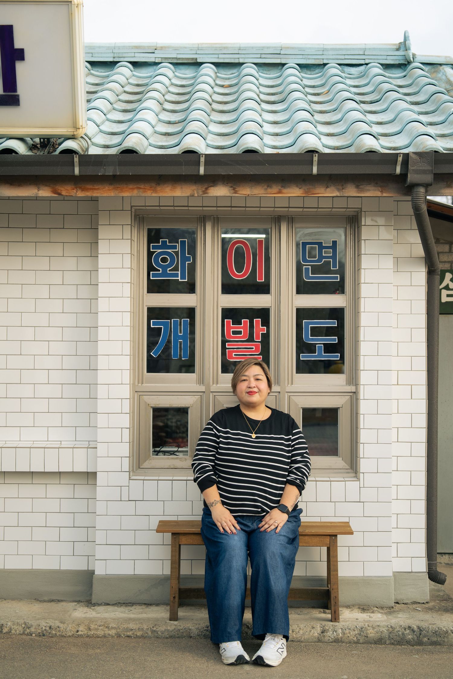 Person sitting on wooden bench outside building with tiled walls and Korean signage.