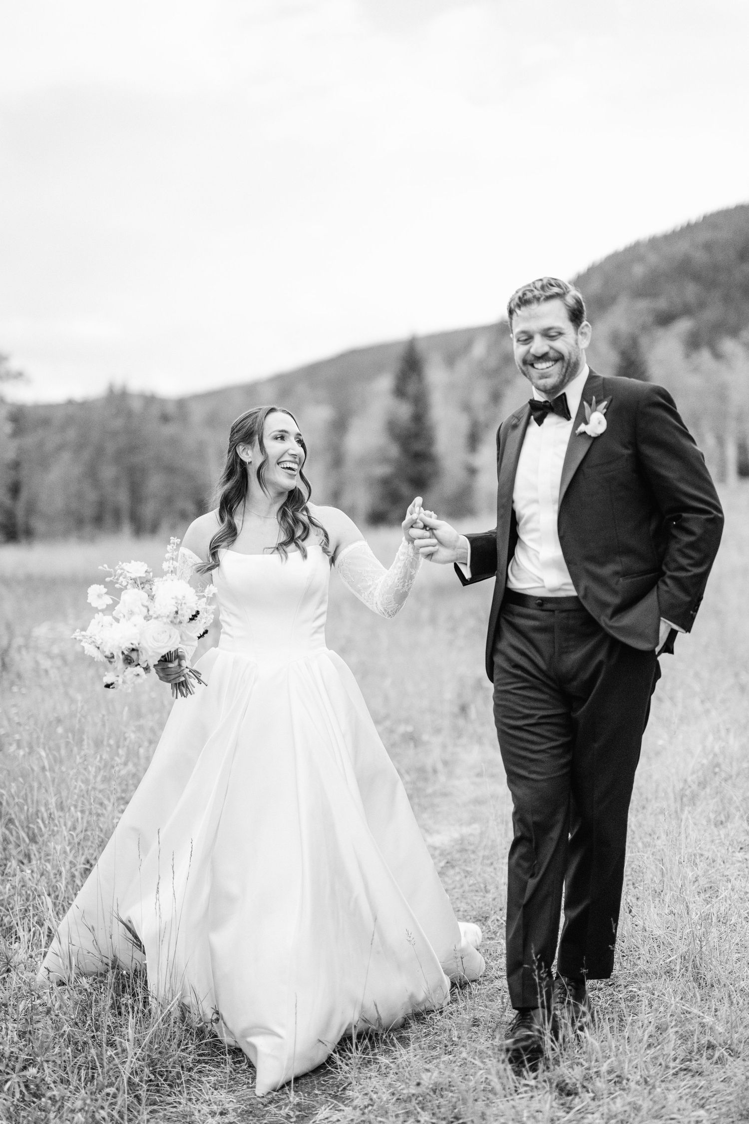 A couple dances joyfully in a mountain meadow with wildflowers and evergreen trees in the background.