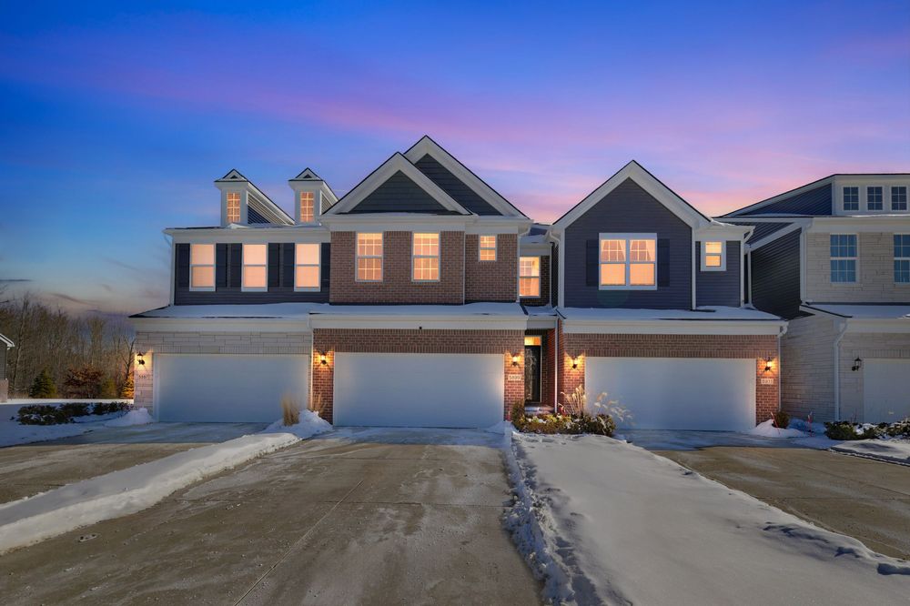 Twilight exterior view of a multi-unit townhome with dark siding, triple garages, and snow-covered driveway.