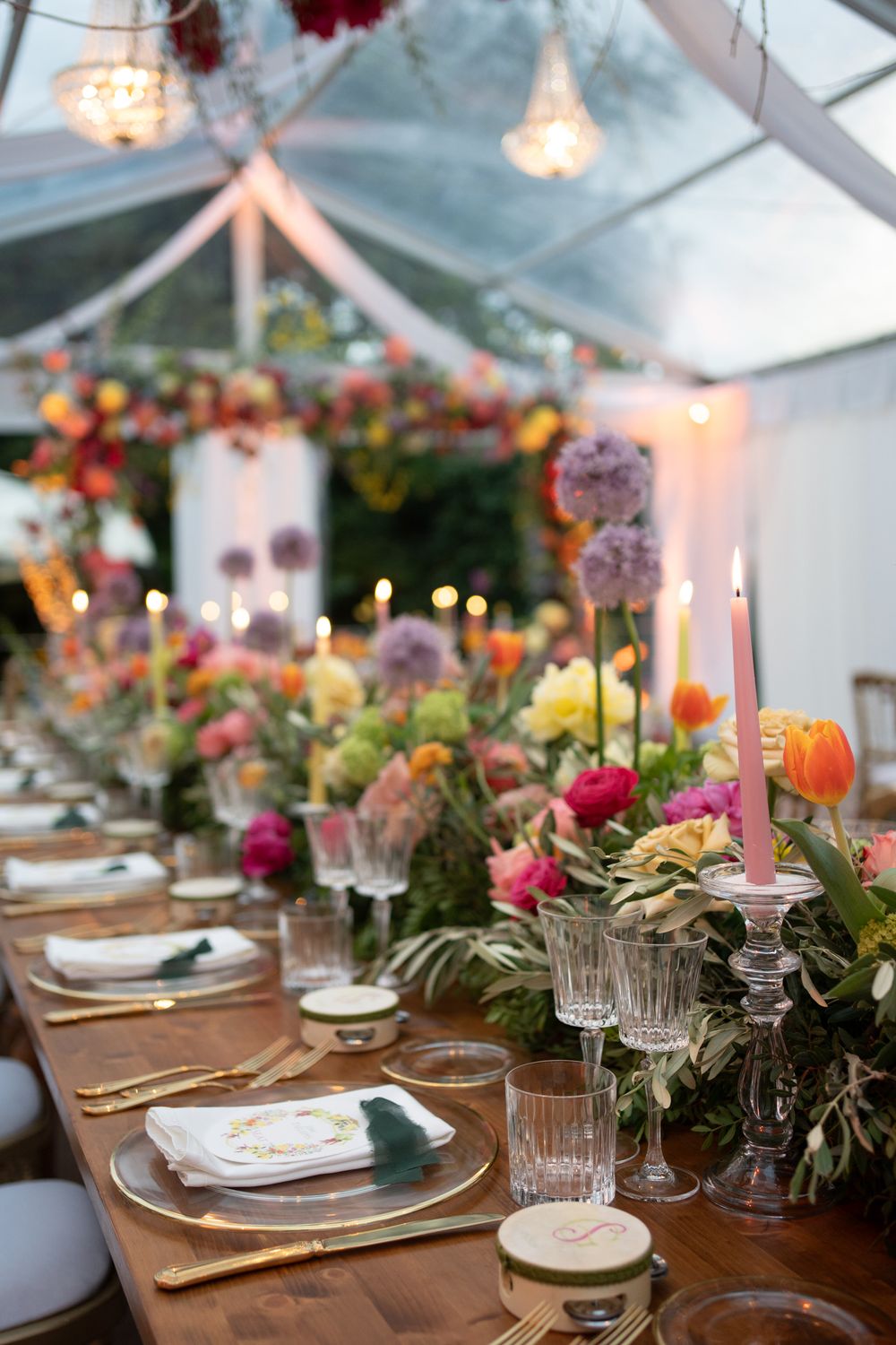 Elegant wedding table setting with pink taper candles and colorful floral centerpieces under a clear tent with chandeliers.