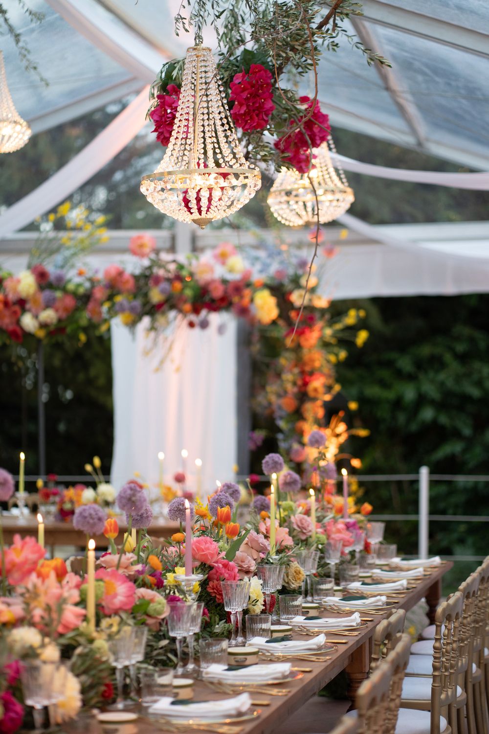 Elegant wedding reception table setup under a clear tent with crystal chandeliers and colorful floral centerpieces.
