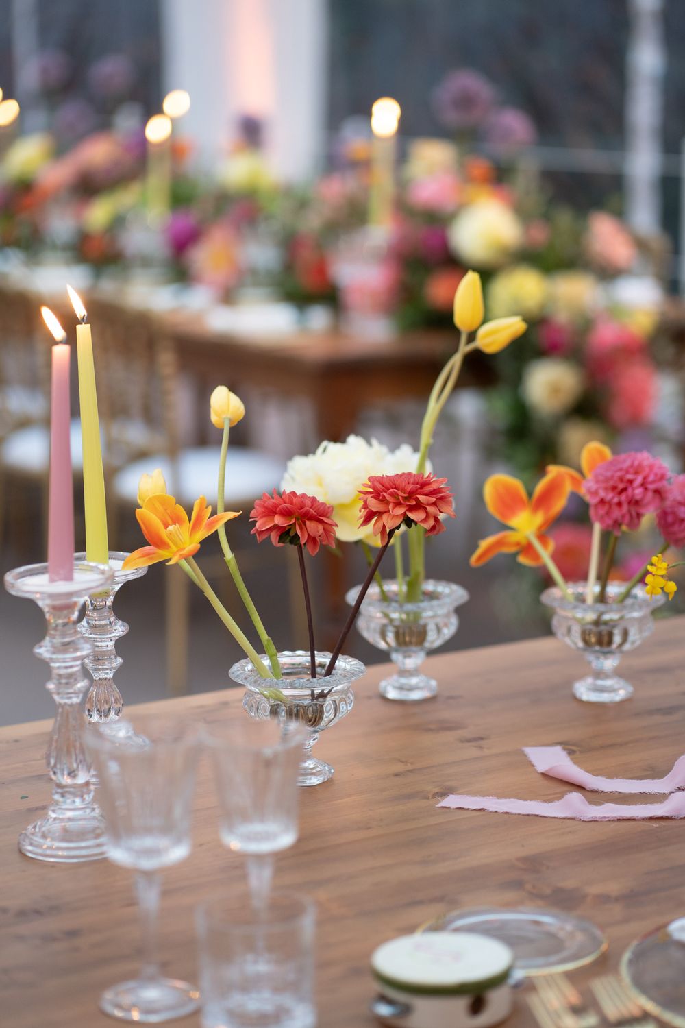 Elegant table setting with small glass vases holding colorful flowers and lit candles on a wooden surface.
