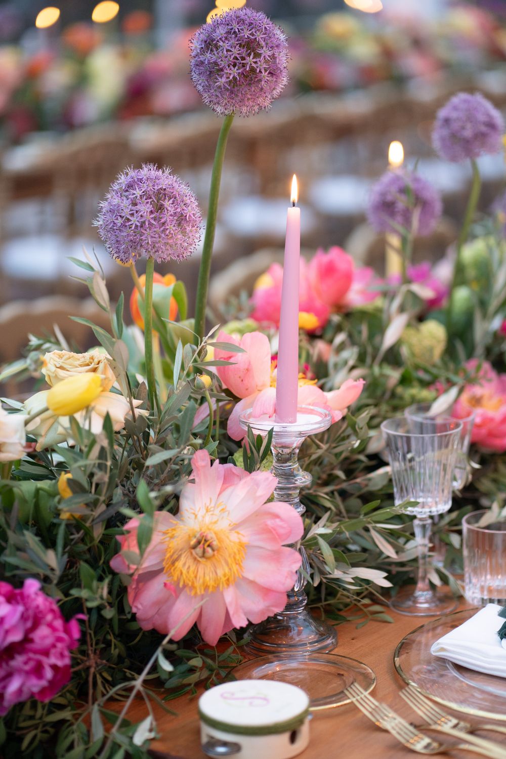 Rustic table setting with pink peonies, purple allium flowers, and pink candles arranged along a wooden surface.