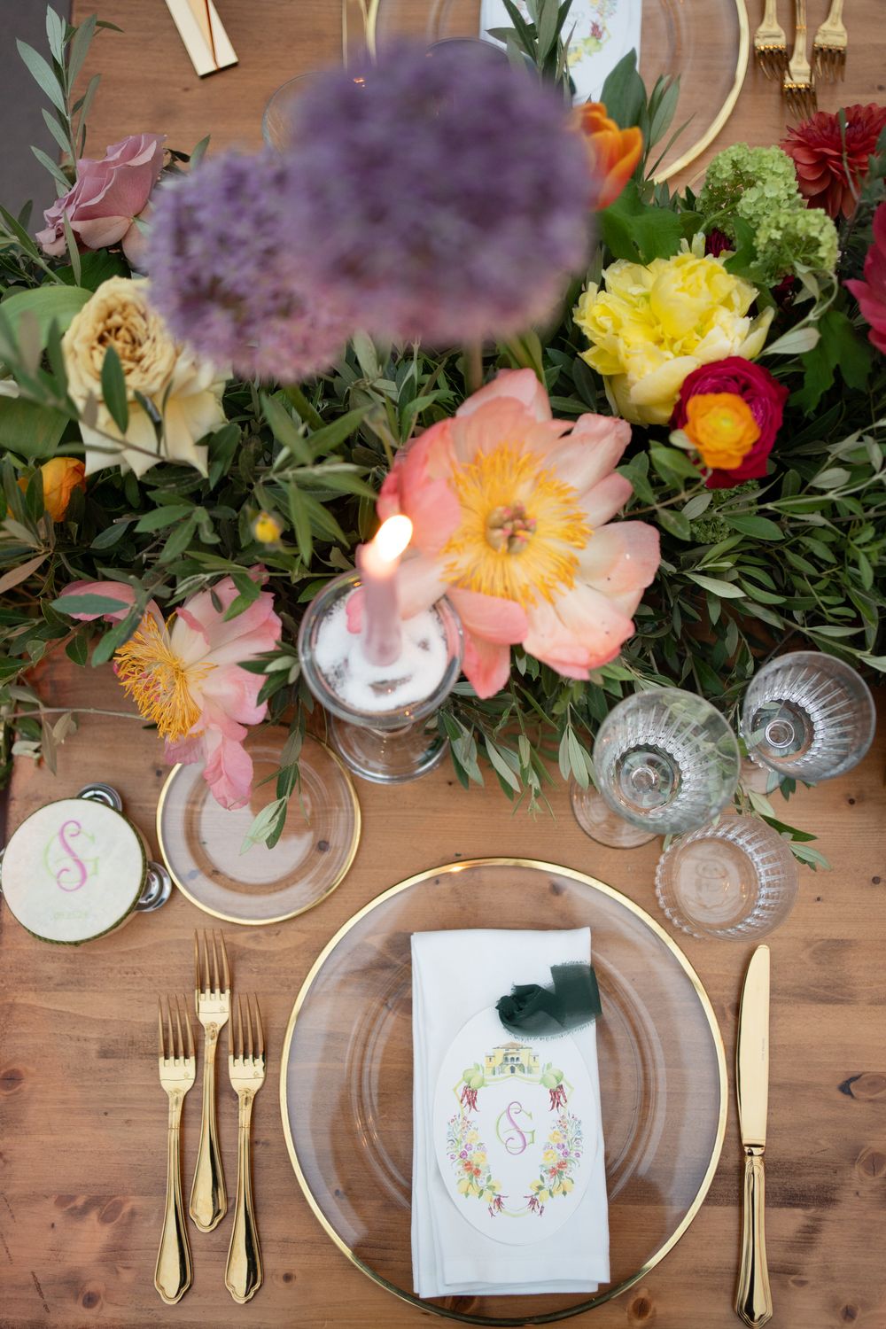 Wedding place setting with gold flatware, crystal glassware, and colorful floral centerpiece on wooden table.