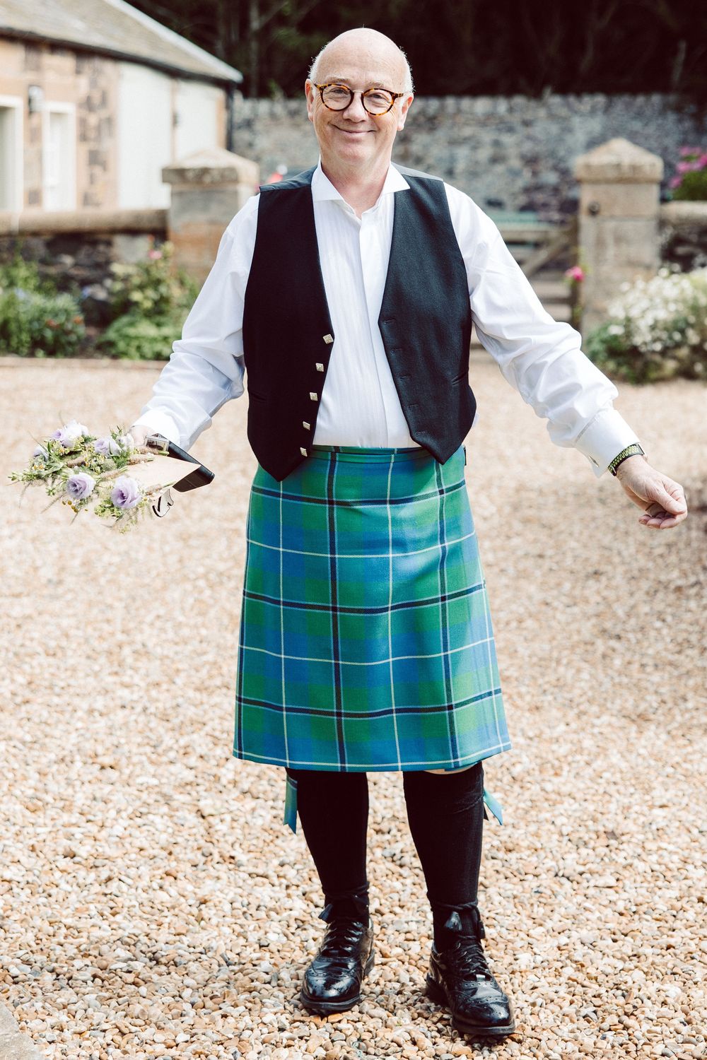 Person wearing a green tartan kilt and black vest poses on gravel path.