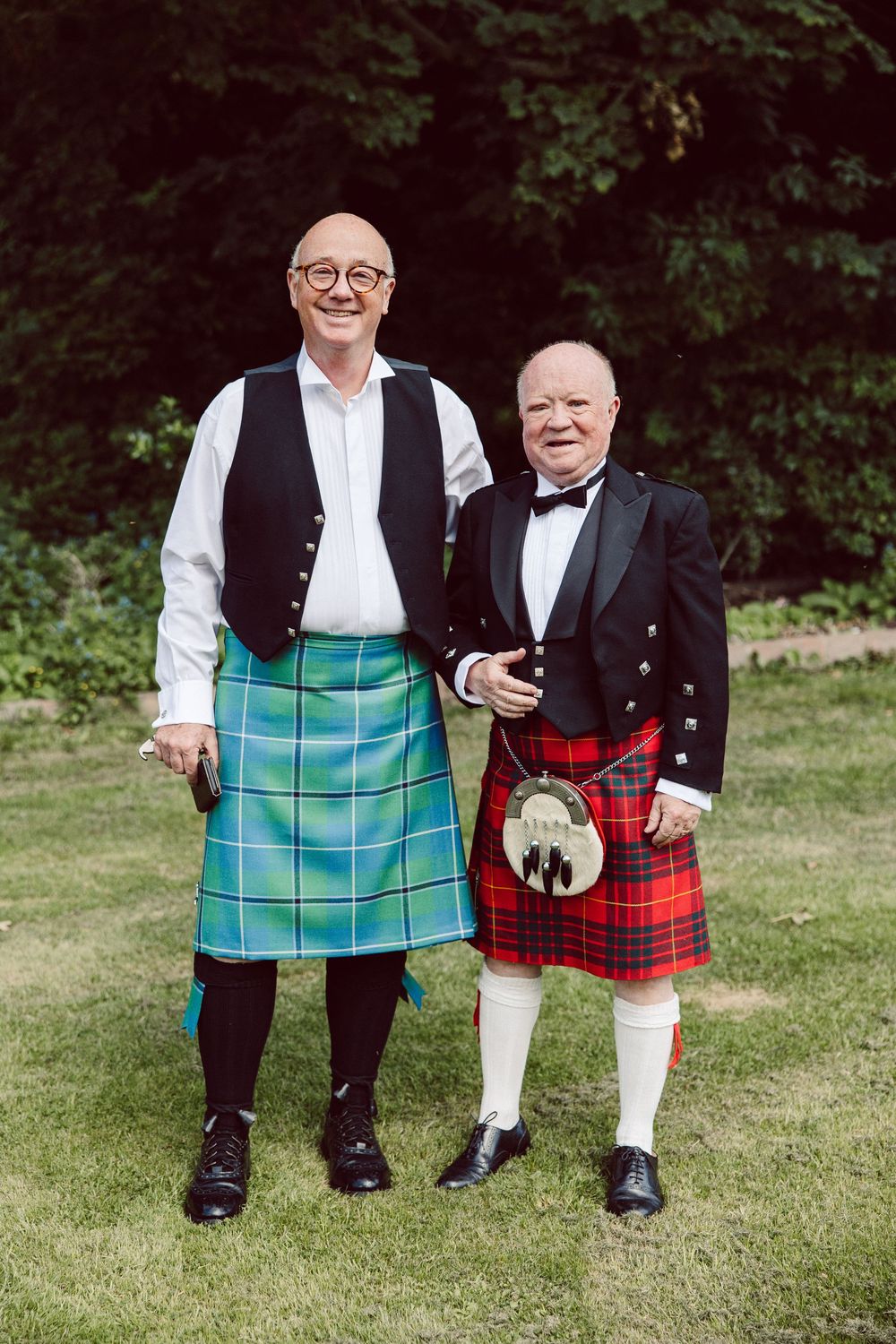 Two men in traditional Scottish kilts and formal attire stand together on grass, one wearing turquoise and one red tartan.
