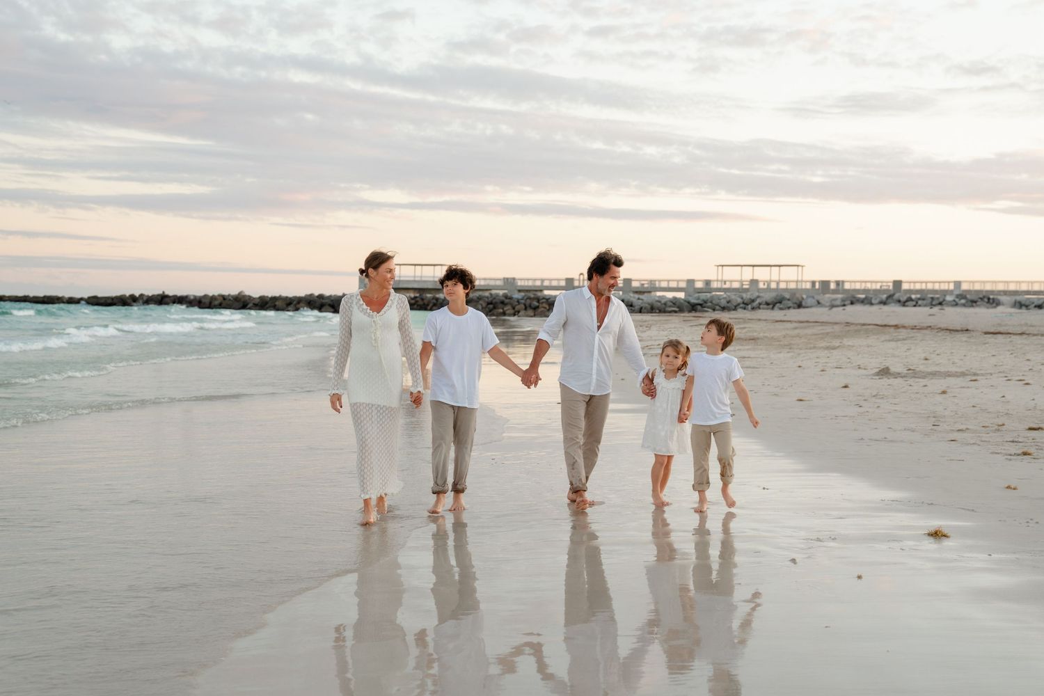 A group in white clothing walks along a sandy beach at sunset creating reflections in the wet sand.
