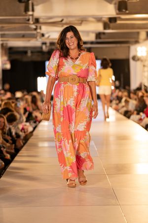 Model walks runway in vibrant orange and pink floral summer dress during fashion show.