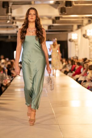 Model walks runway in flowy sage green dress during fashion show event.