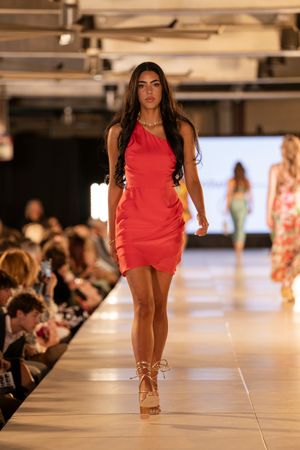 Models present vibrant red summer outfits on white runway with audience in background.