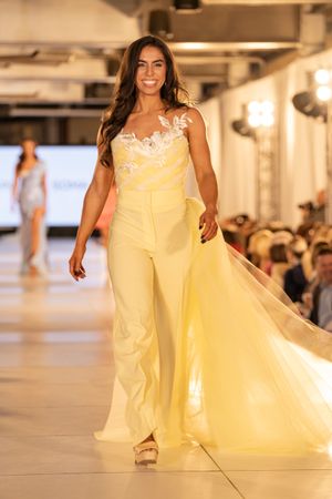 Model walks runway in flowing cream colored wedding dress with dramatic cape detail during bridal fashion show.