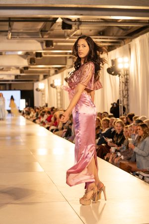 Model showcases flowing pink satin evening gown on illuminated catwalk with audience.