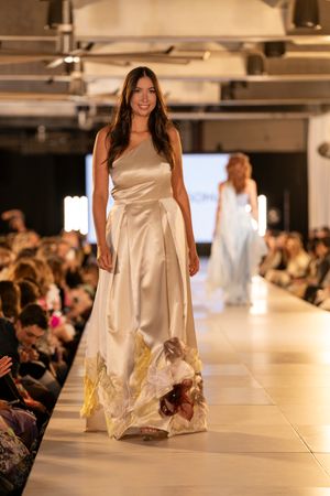Model showcases an elegant white satin gown on a well-lit fashion show runway with audience in background.