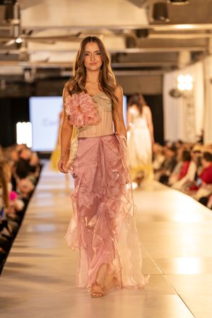 Models showcase flowing pink ruffled dresses on an illuminated fashion runway with audience seated in background.