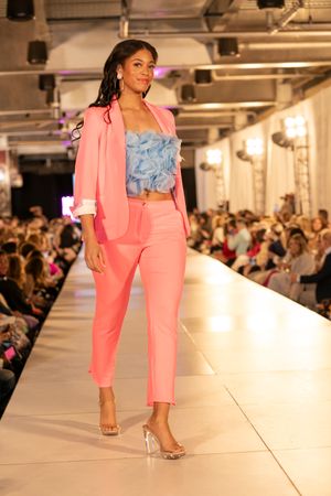 Model walks the runway in a coral pink pantsuit and silver heels during a fashion show event.