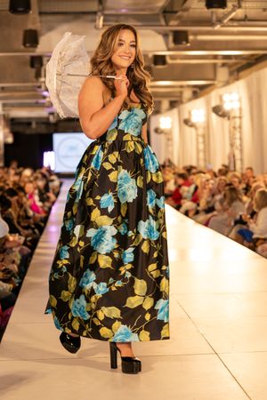 Fashion model walks runway in floral print dress with blue and green pattern during indoor fashion show.