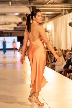 Model walks the runway in a flowing peach-colored dress and nude heels at a fashion show.
