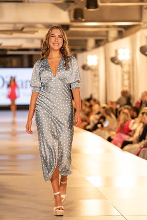 Model walks runway in polka dot gray silk wrap dress with puff sleeves and white platform sandals at fashion show.