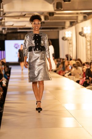 Model struts confidently in polka dot mini dress on brightly lit runway.
