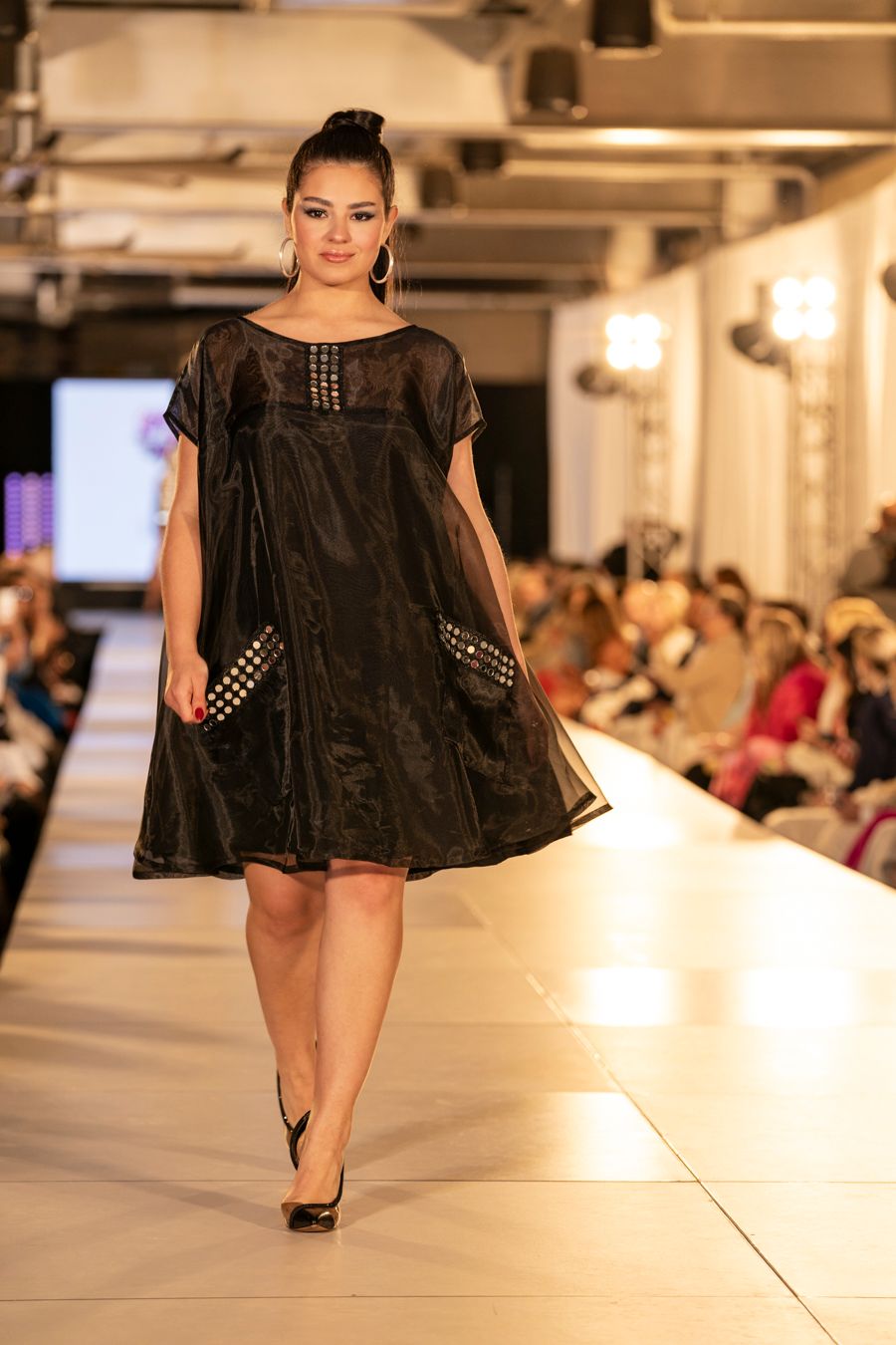 Model walks runway in black sheer cape with studded details during fashion show.
