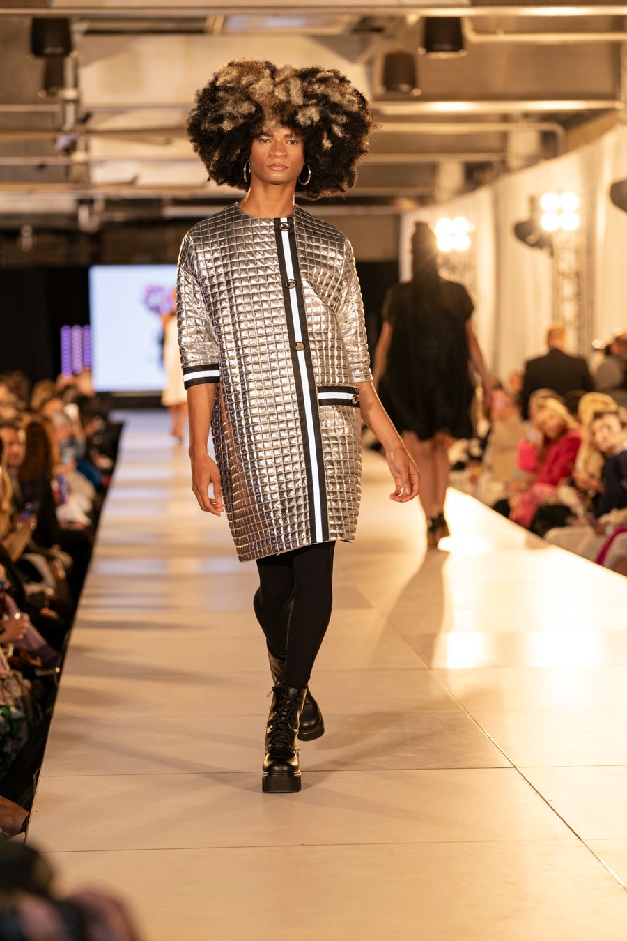 Model walks runway in silver metallic checkered jacket and black pants during fashion show.