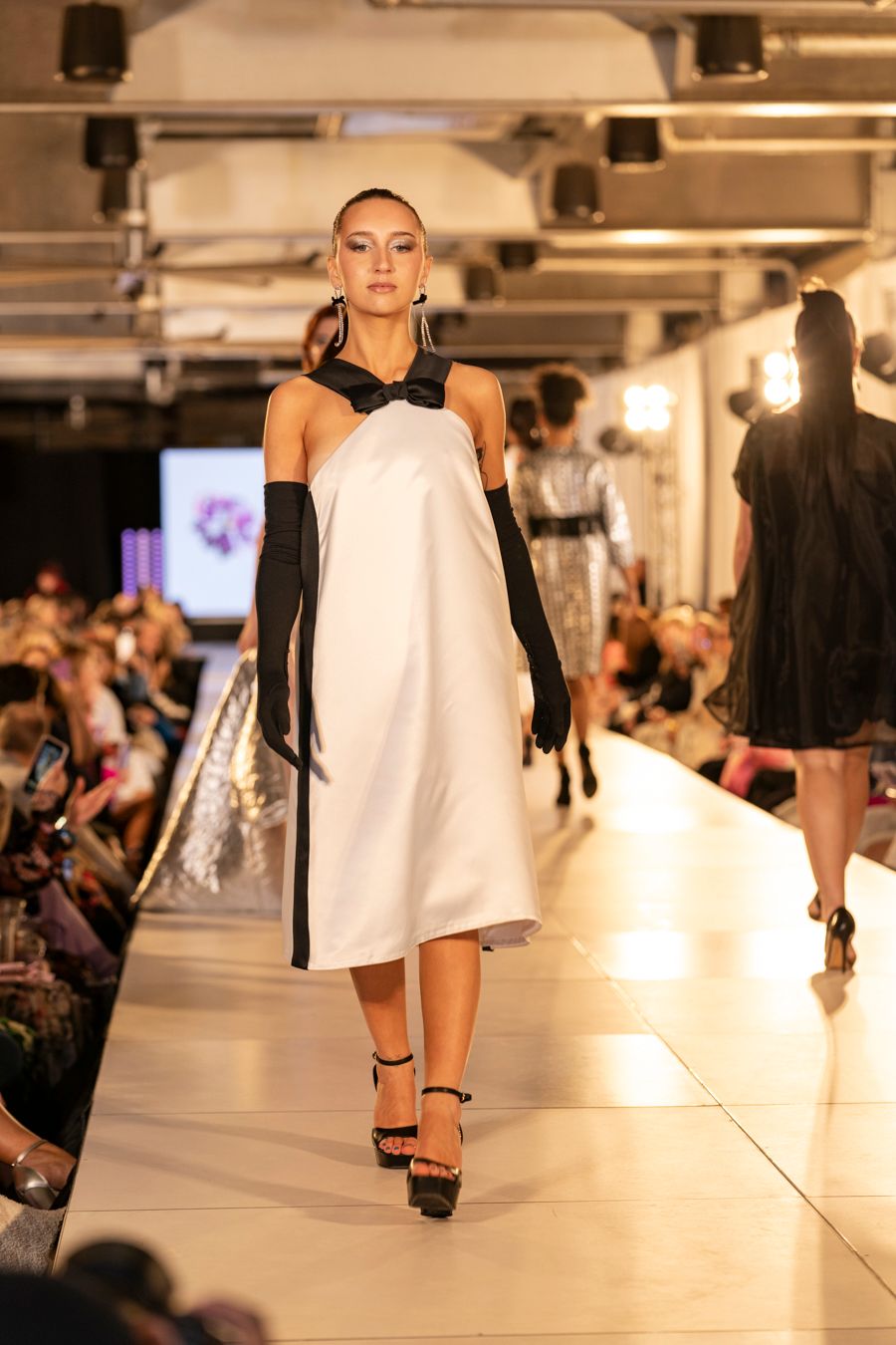Models walk the runway in white asymmetrical dresses with black accessories during a fashion show.