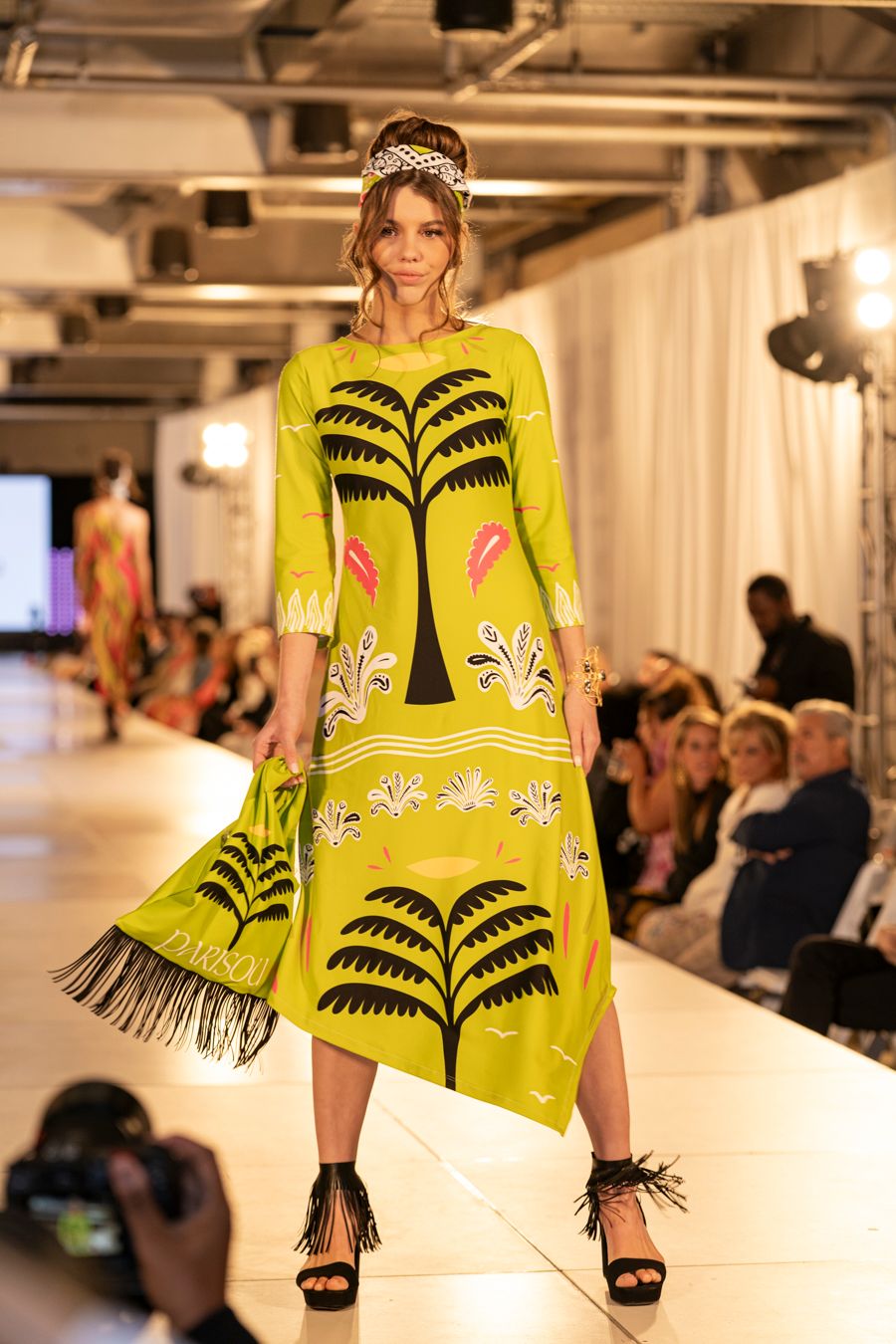 Fashion model walks runway in bright yellow dress with black palm tree pattern and fringe details at designer show.