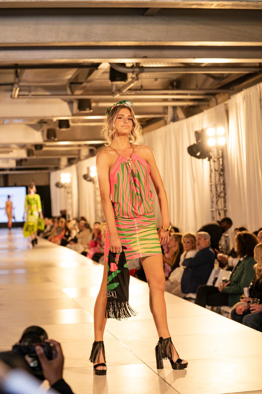 Fashion model walks runway in neon green swimwear during fashion show with audience seated along catwalk.