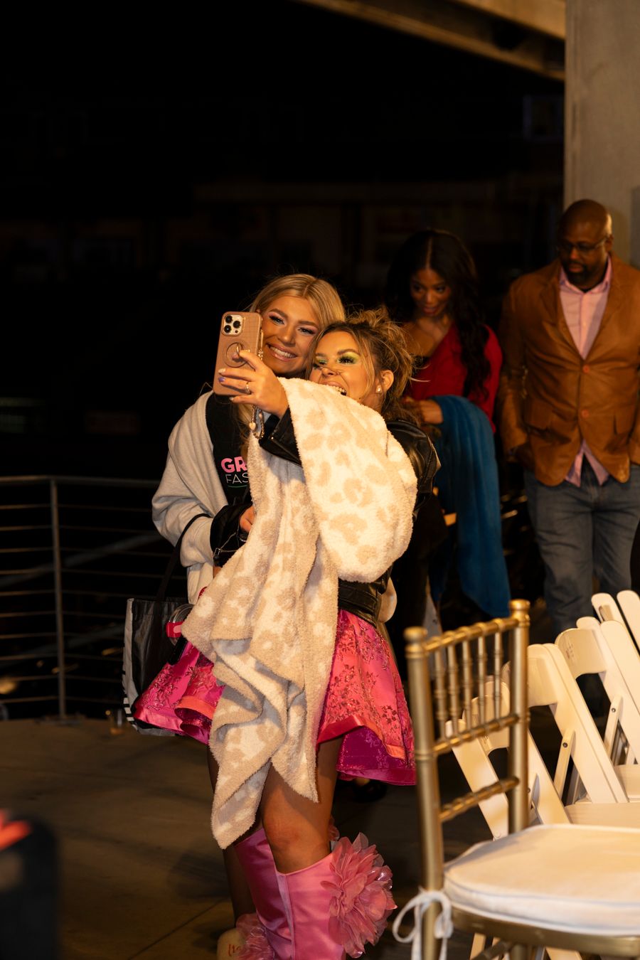 Two people take a selfie together at an event with white chairs and decorative lighting in the background.